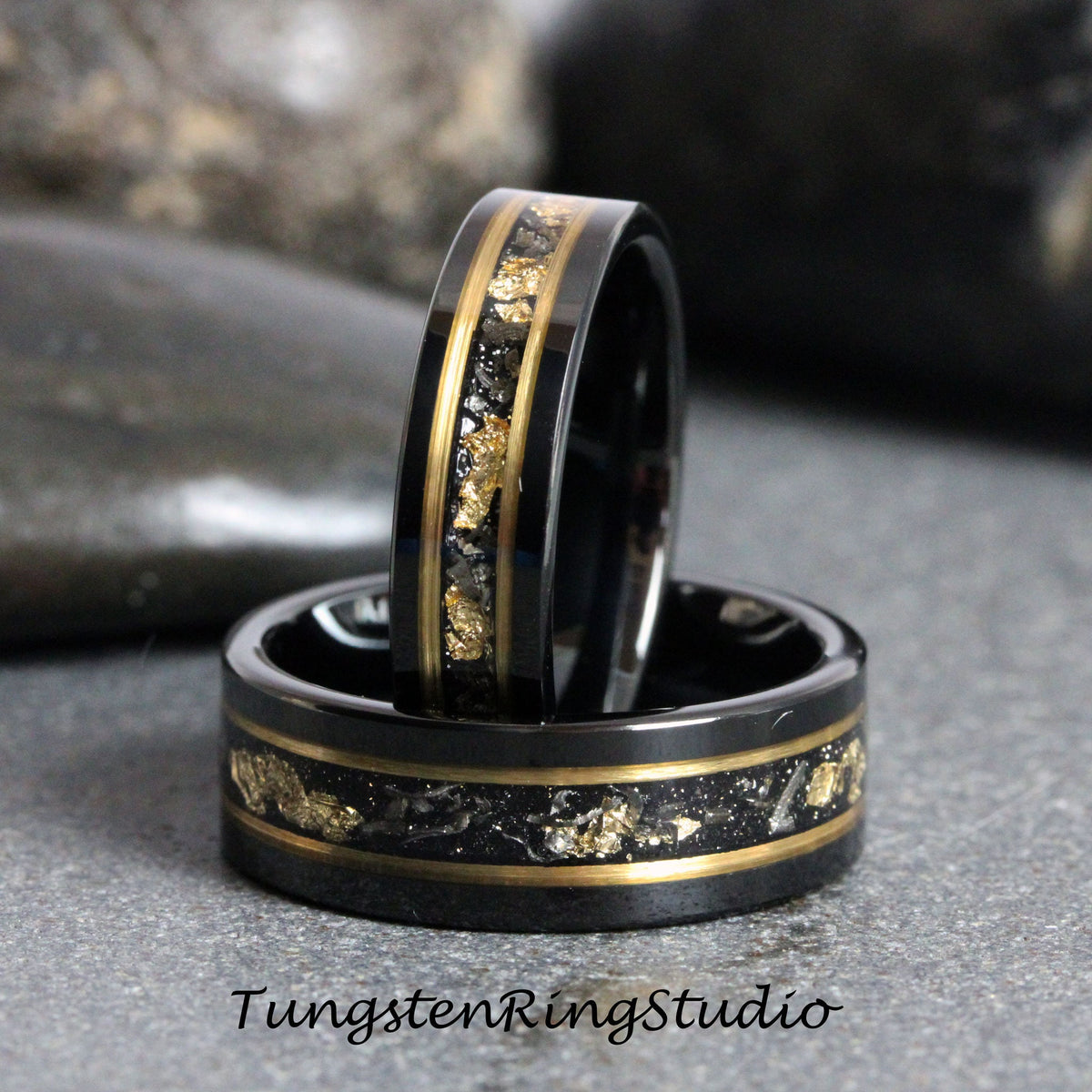 May include: Two black tungsten carbide rings with gold inlay and black specks. The rings are stacked on top of each other.