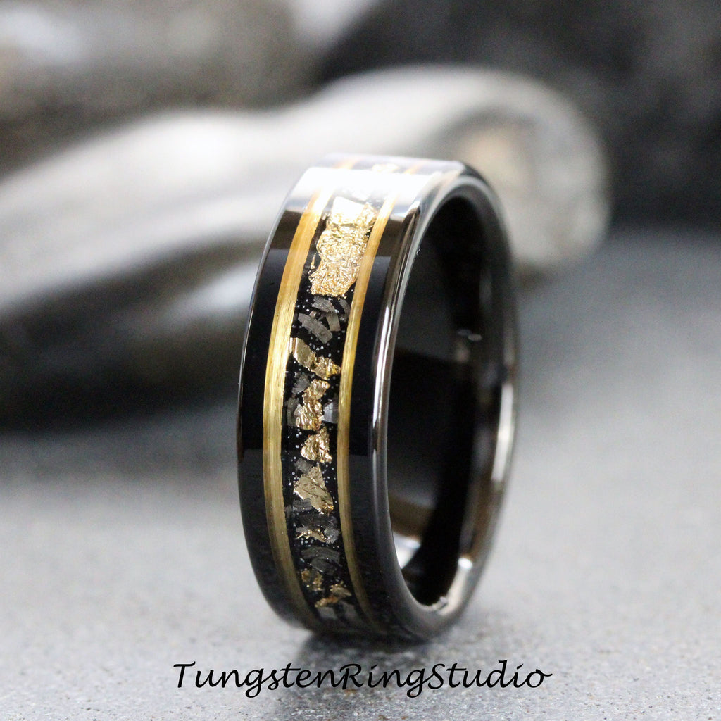 May include: A black tungsten carbide ring with a gold inlay featuring a pattern of small gold flakes. The ring has a polished finish.