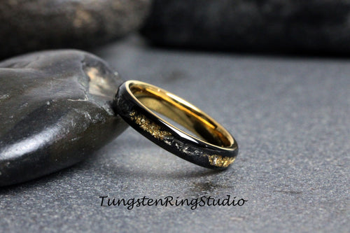 May include: A black tungsten ring with a gold inlay. The inlay is a thin line of gold that runs around the entire ring. The ring is on a gray surface.