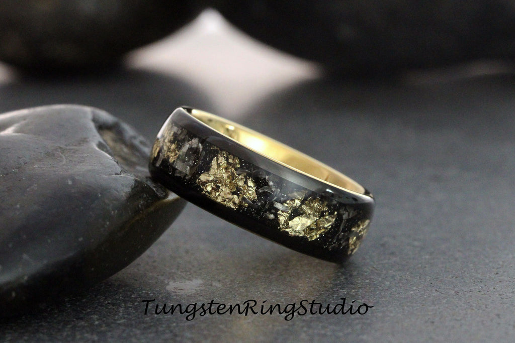 May include: A black tungsten ring with a gold inlay featuring a design of gold flakes embedded in black resin. The ring is shown on a dark gray surface.