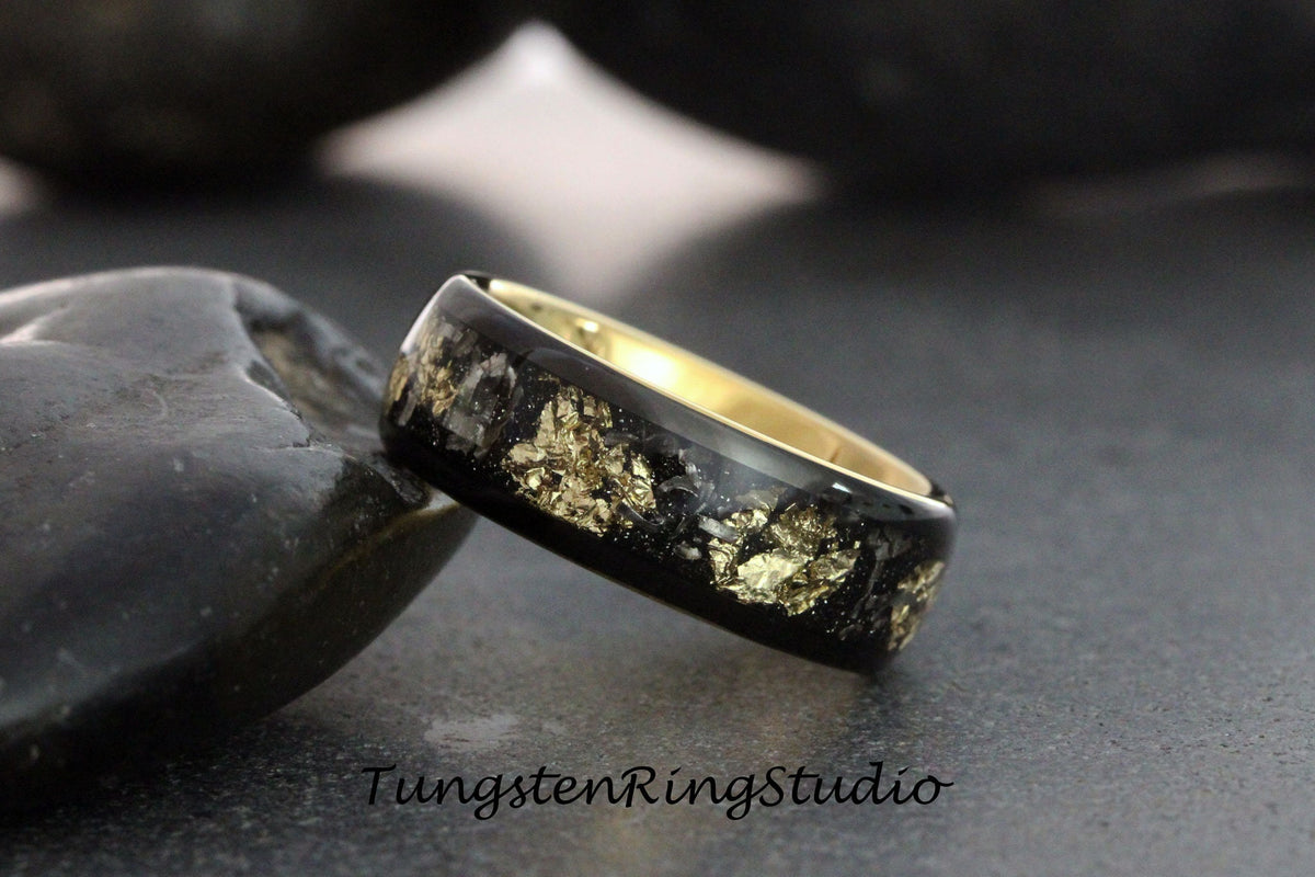 May include: A black tungsten ring with a gold inlay featuring a design of gold flakes embedded in black resin. The ring is shown on a dark gray surface.
