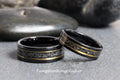 May include: Two black tungsten carbide rings with a gold inlay and a black speckled design. Tungsten Ring Studio is printed below the rings.