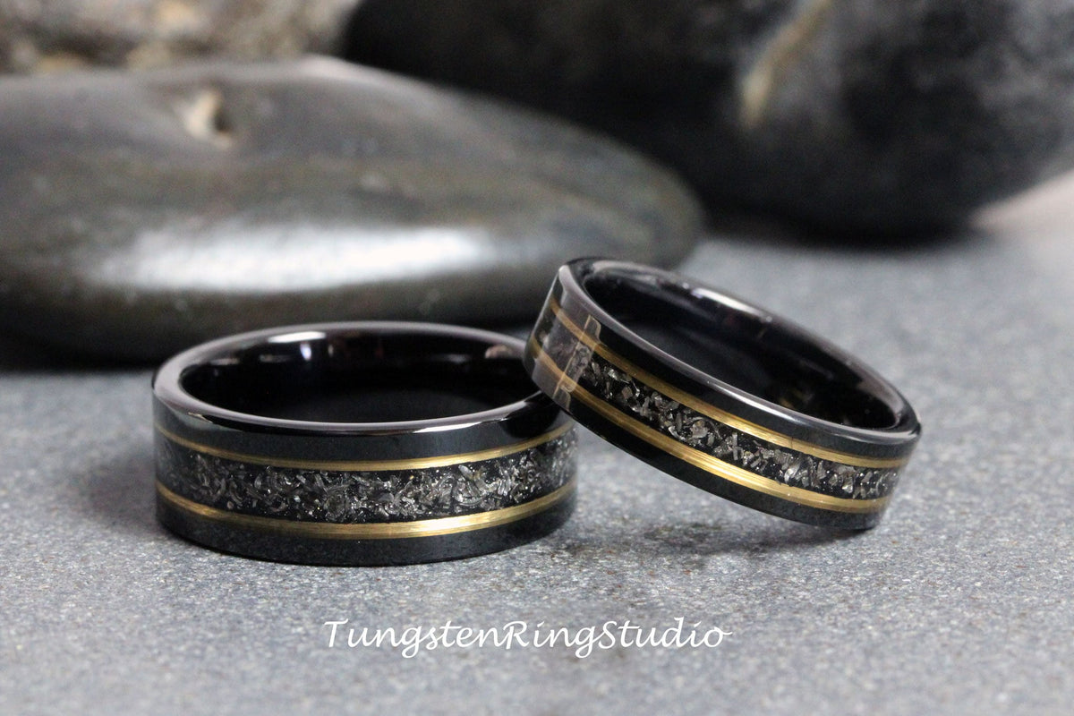May include: Two black tungsten carbide rings with a gold inlay and a black speckled design. Tungsten Ring Studio is printed below the rings.