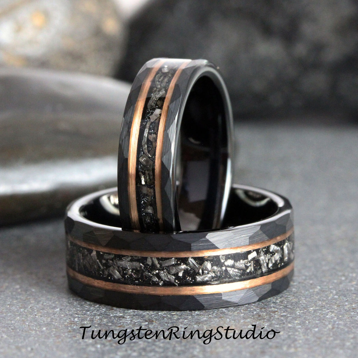 May include: Two black tungsten carbide rings with a hammered finish and a rose gold inlay. The inlay features a pattern of small, silver-colored particles.