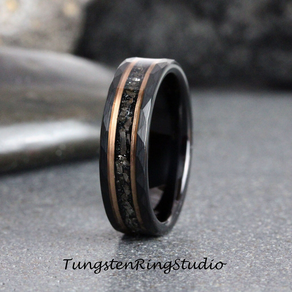 May include: A black tungsten carbide ring with a beveled edge and a rose gold inlay featuring black particles. The ring is on a gray surface.