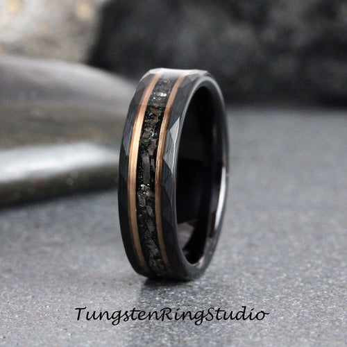 May include: A black tungsten carbide ring with a beveled edge and a rose gold inlay featuring black particles. The ring is on a gray surface.