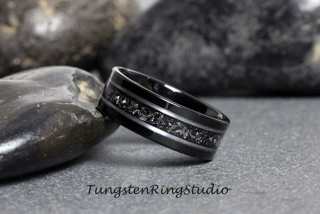 May include: A black tungsten carbide ring with a silver inlay design. The ring has a brushed finish and a beveled edge.