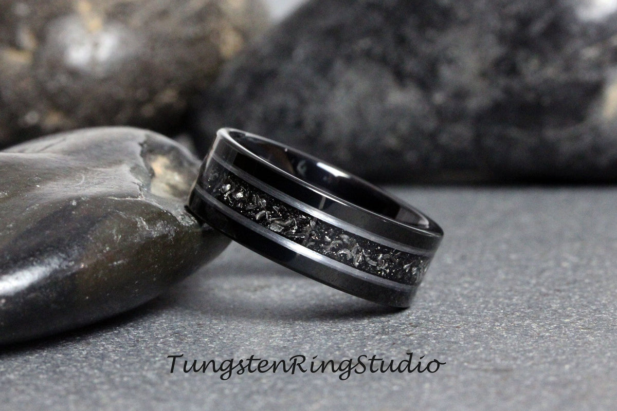 May include: A black tungsten carbide ring with a silver inlay design. The ring has a brushed finish and a beveled edge.