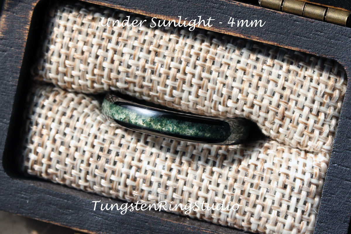 May include: A close-up of a dark gray tungsten ring with a green and black inlay. The ring is sitting on a beige fabric background. The text "Under Sunlight - 4mm" is visible above the ring. The text "TungstenRingStudio" is visible below the ring.