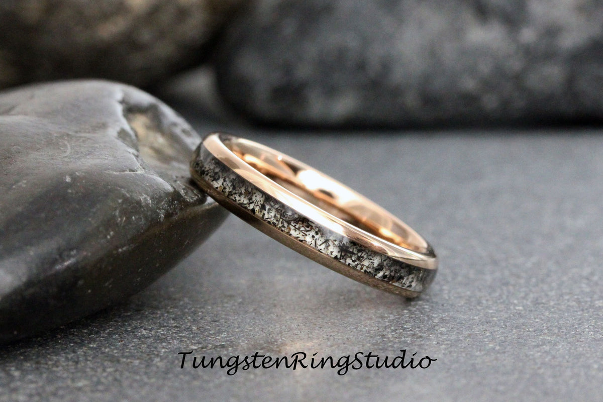 May include: A close-up of a rose gold tungsten carbide ring with a black and gray inlay. The ring is sitting on a gray rock.