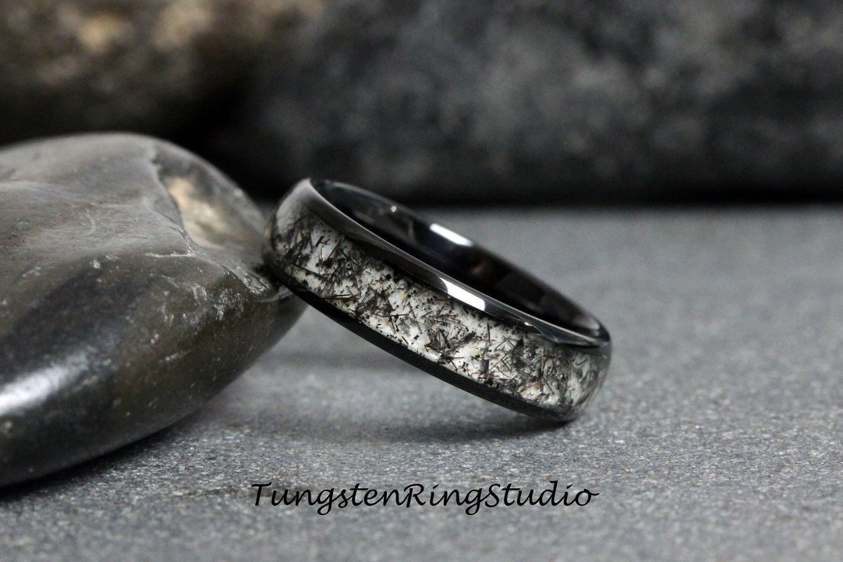 May include: A black tungsten carbide ring with a white inlay. The inlay has a unique, speckled pattern. Tungsten Rings Studio is printed on the surface below the ring.