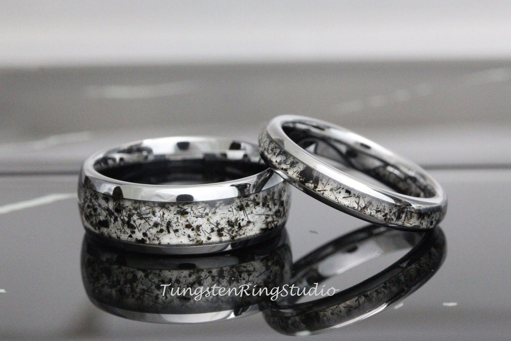 May include: A pair of silver tungsten carbide rings with a black and white speckled inlay. The rings are shown on a reflective surface.