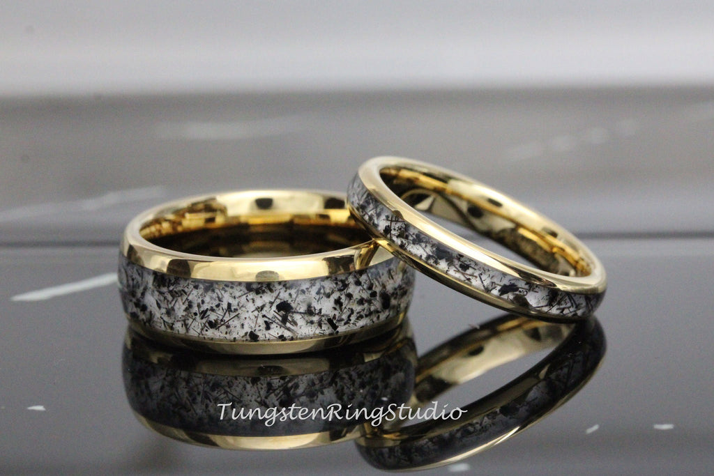 May include: A pair of tungsten carbide wedding bands with a gold inlay and a black and white speckled design.  The bands are shown on a black surface.