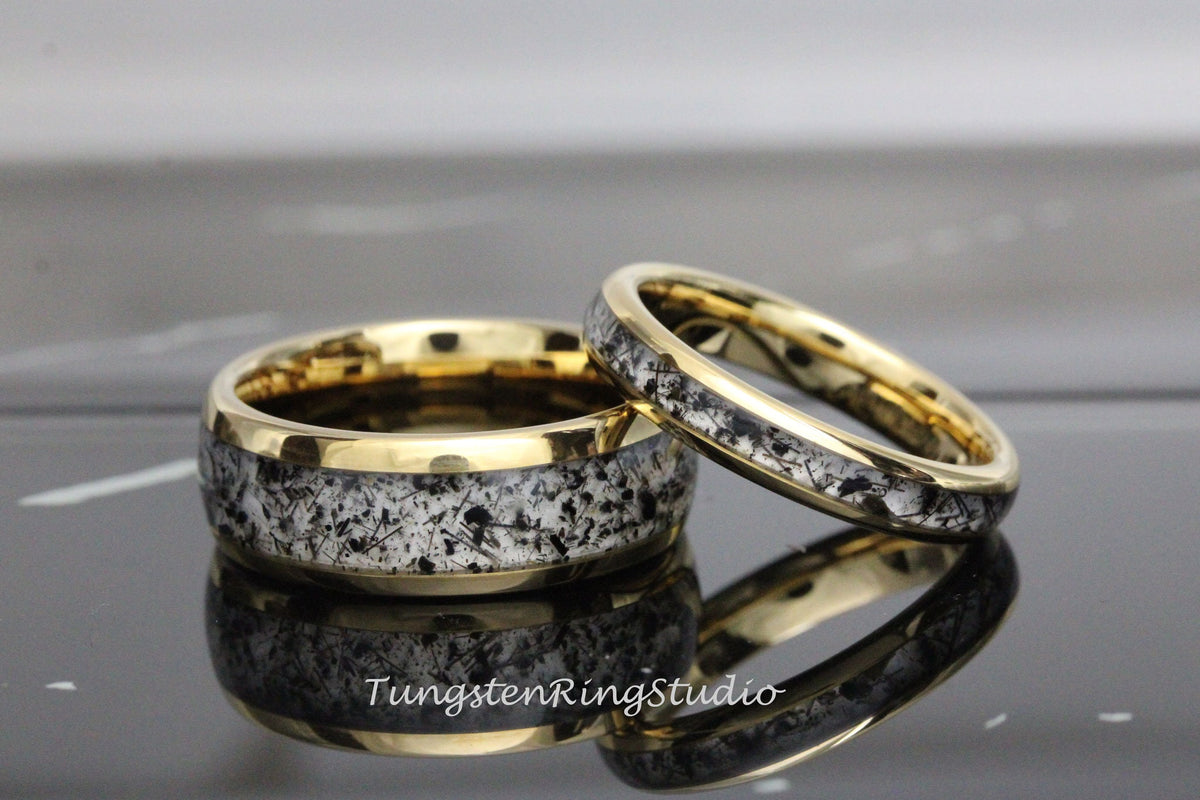 May include: A pair of tungsten carbide wedding bands with a gold inlay and a black and white speckled design.  The bands are shown on a black surface.