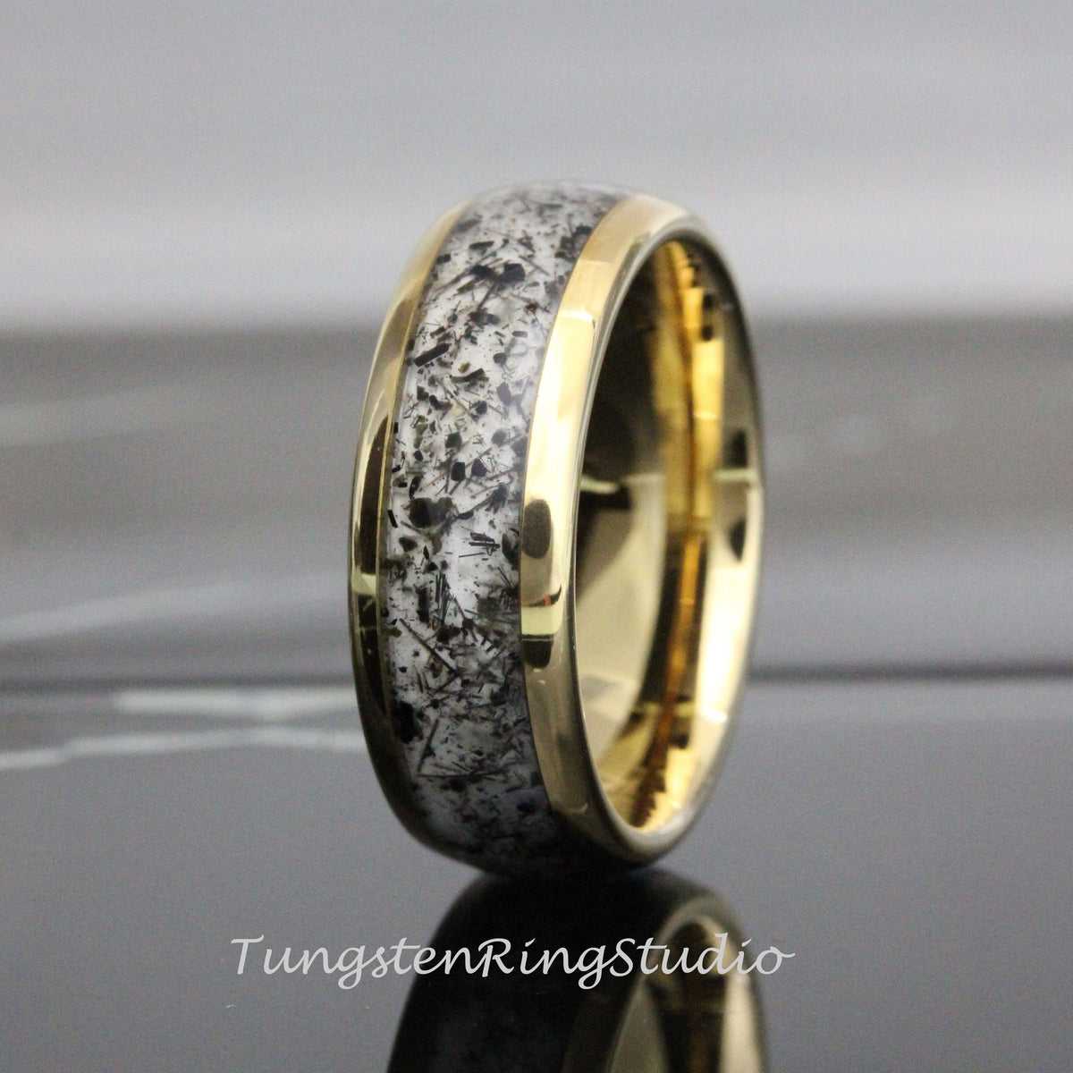 May include: A gold-colored ring with a band of white and black speckled material. The ring has a polished finish and a smooth, rounded shape. The text "TungstenRingStudio" is visible at the bottom of the image.
