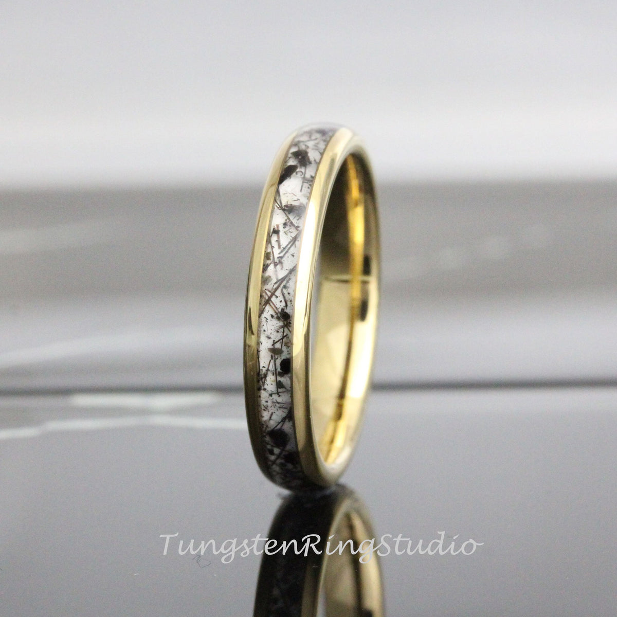 May include: A gold-colored ring with a band of white and black speckled material. The ring has a polished, reflective surface. The text "TungstenRingStudio" is visible at the bottom of the image.