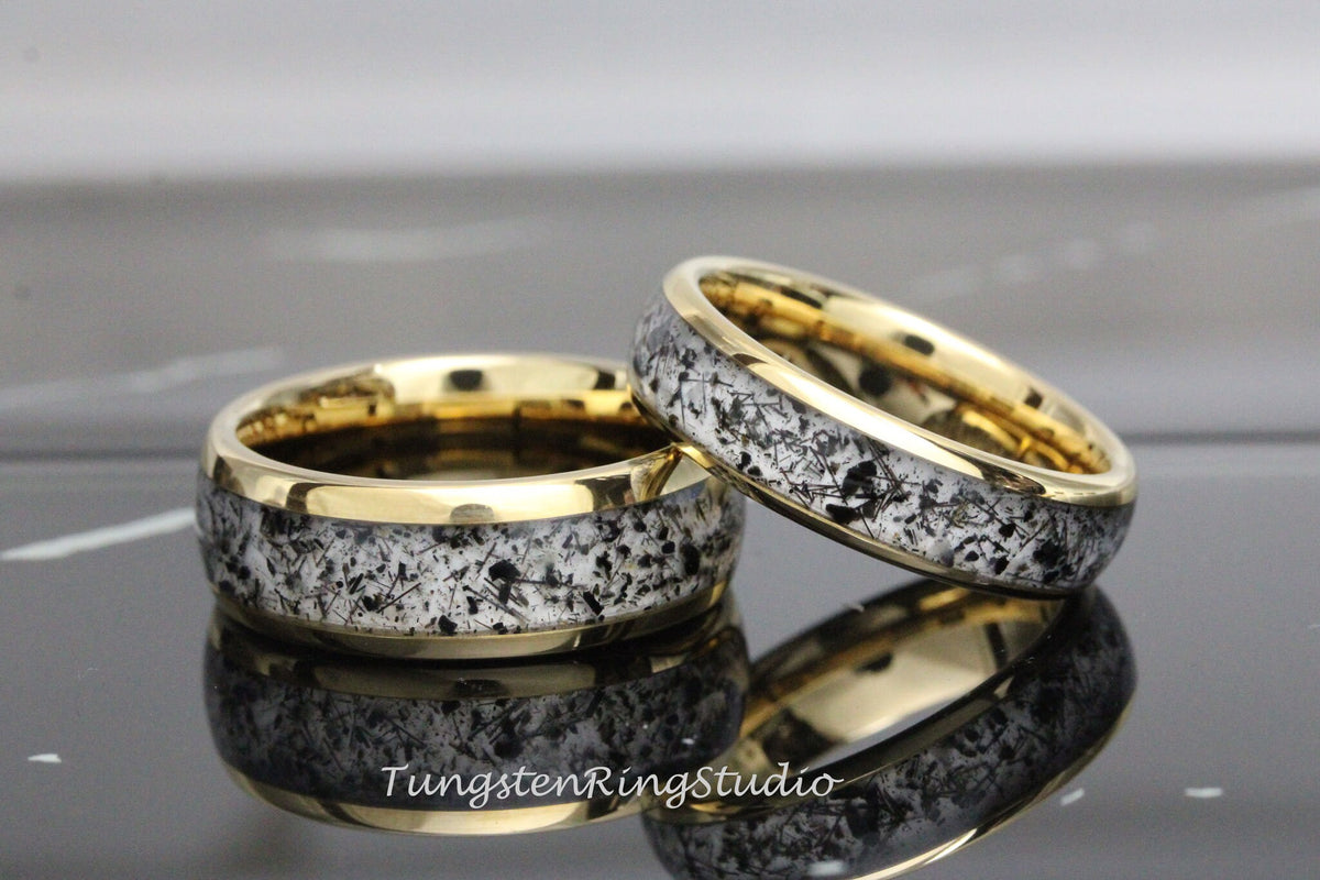 May include: Two gold-plated rings with a unique design. The rings feature a band of speckled black and white material set within a gold frame. The rings are displayed on a reflective surface.