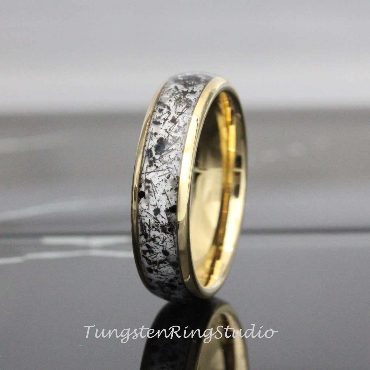 May include: A gold-colored ring with a band featuring a white and black speckled design. The ring has a polished finish and a smooth, rounded profile. The ring is a piece of jewelry.