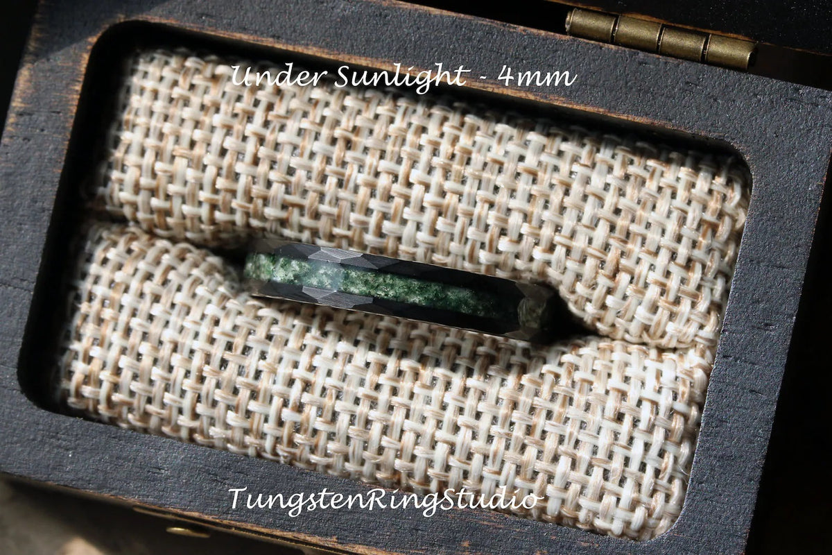 May include: A black tungsten carbide ring with a green inlay, displayed in a wooden box with a burlap lining. The ring is 4mm wide and the text "Under Sunlight - 4mm" is visible above the ring. The text "TungstenRingStudio" is visible below the ring.
