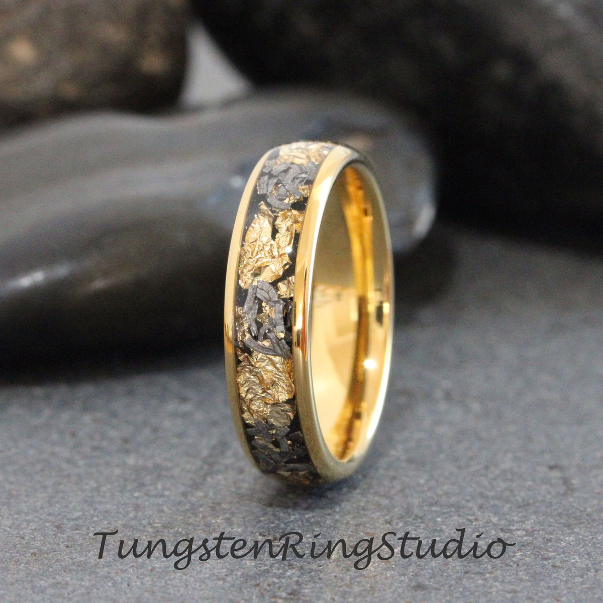May include: A gold and black tungsten carbide ring with a band of gold and a black inlay with gold flecks. The ring is on a gray background.