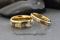 May include: Two gold tungsten carbide wedding bands with a black and gold inlay. The bands are on a gray background.