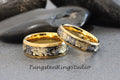 May include: Two gold tungsten carbide rings with a black and gold inlay. The rings are on a gray background.