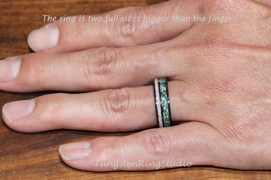 May include: A black tungsten ring with a green and pink inlay. The ring is worn on a finger. The ring is two full sizes bigger than the finger. The text "TungstenRingStudio" is visible in the image.