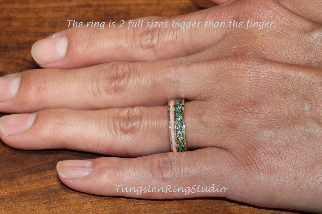 May include: A close-up of a hand wearing a tungsten ring with a green and pink inlay. The ring is two sizes larger than the finger. The text "TungstenRingStudio" is visible in the lower right corner of the image.