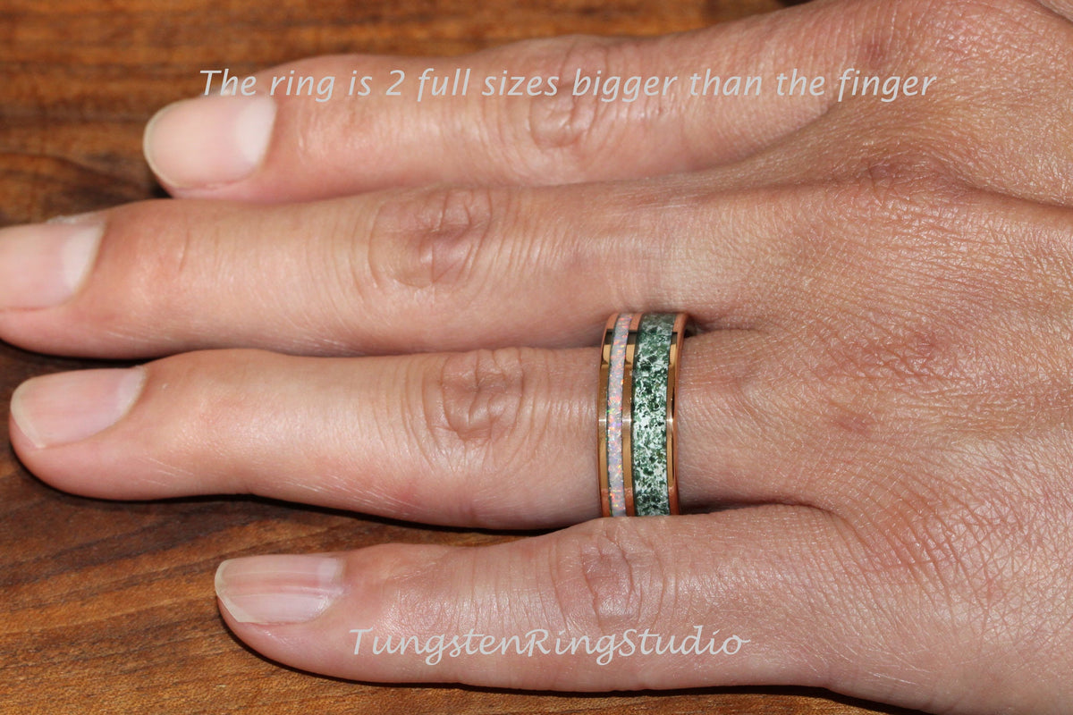 May include: A close-up of a hand wearing a green and pink tungsten ring with a gold band. The ring is two sizes bigger than the finger. The text "TungstenRingStudio" is visible in the bottom right corner of the image.