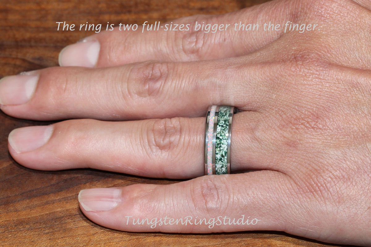May include: A close-up of a hand wearing a tungsten ring with a green and pink inlay. The ring is two full sizes bigger than the finger. The text "TungstenRingStudio" is visible in the bottom right corner of the image.