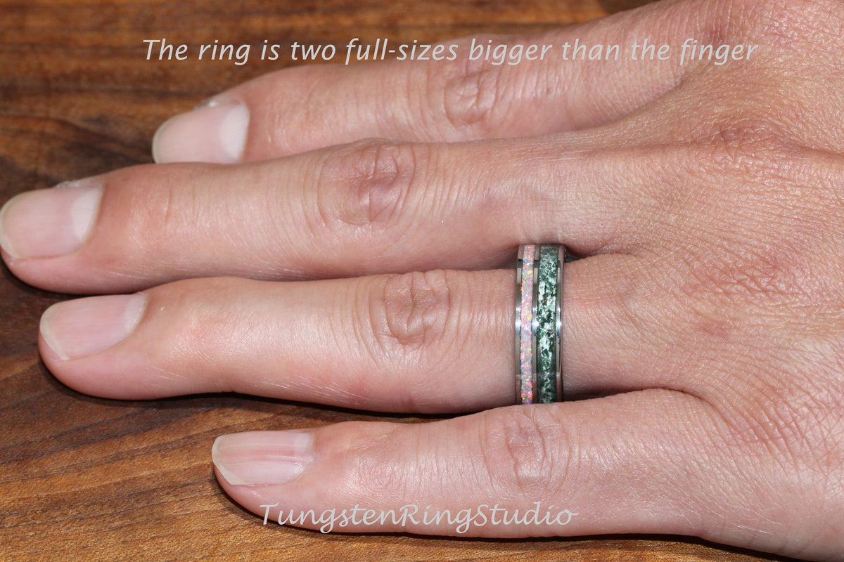 May include: A close-up of a hand wearing a tungsten ring with a green and pink inlay. The ring is two sizes larger than the finger. The text "TungstenRingStudio" is visible in the lower right corner of the image.
