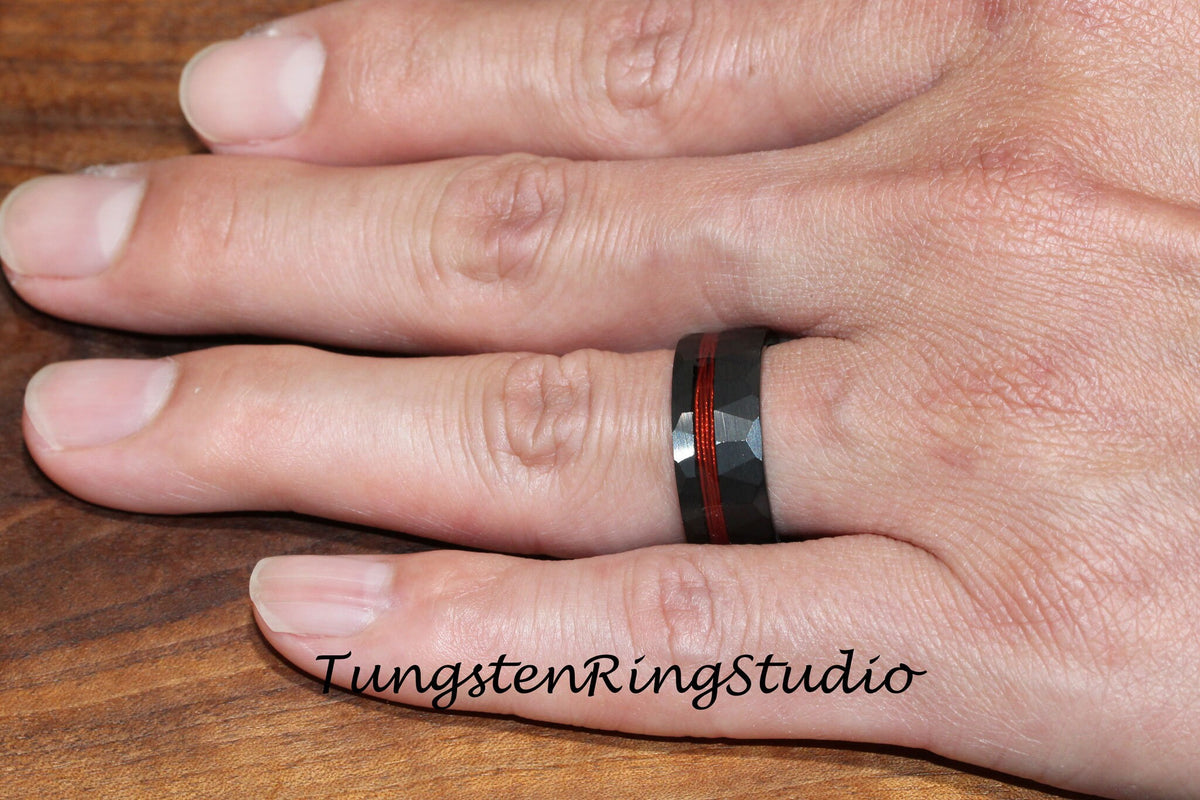 May include: A black tungsten carbide ring with a red inlay and a hammered finish. The ring is worn on a person's finger.