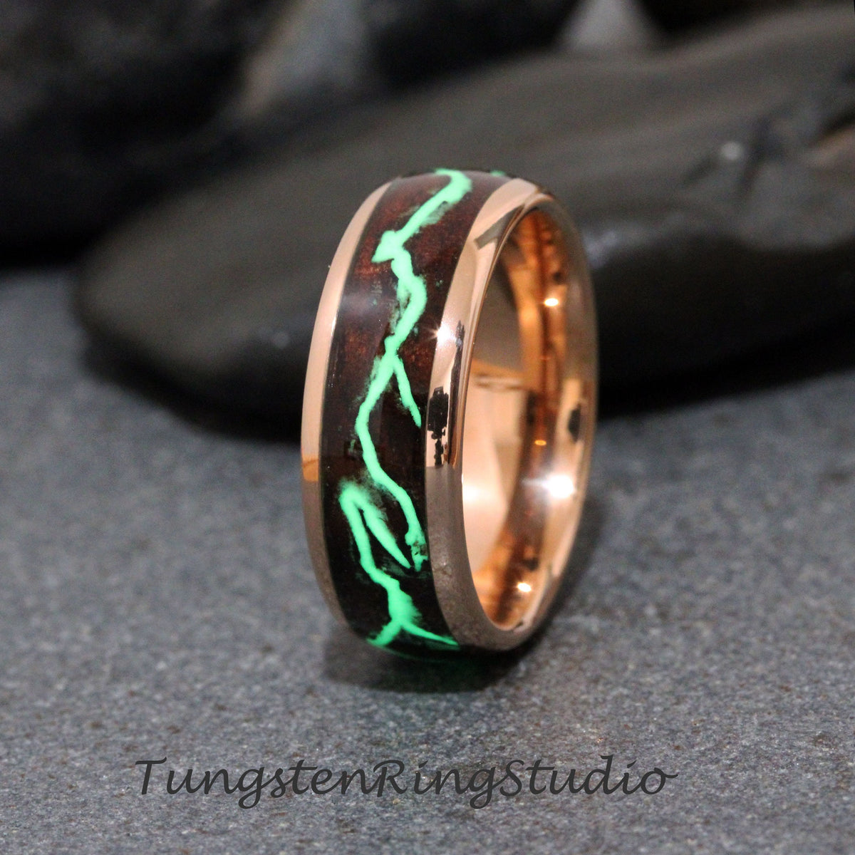 May include: A rose gold tungsten carbide ring with a dark brown wood inlay and a green glow-in-the-dark design. The ring is a men's wedding band.