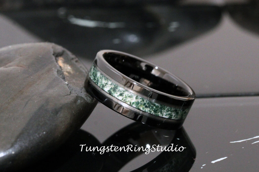 May include: A black tungsten ring with a green and white inlay. The ring has a polished finish and a beveled edge. Tungsten Ring Studio is printed on the surface below the ring.