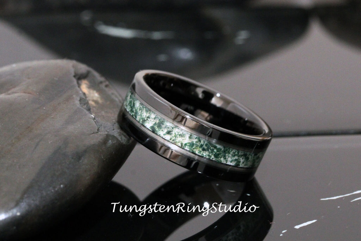 May include: A black tungsten ring with a green and white inlay. The ring has a polished finish and a beveled edge. Tungsten Ring Studio is printed on the surface below the ring.