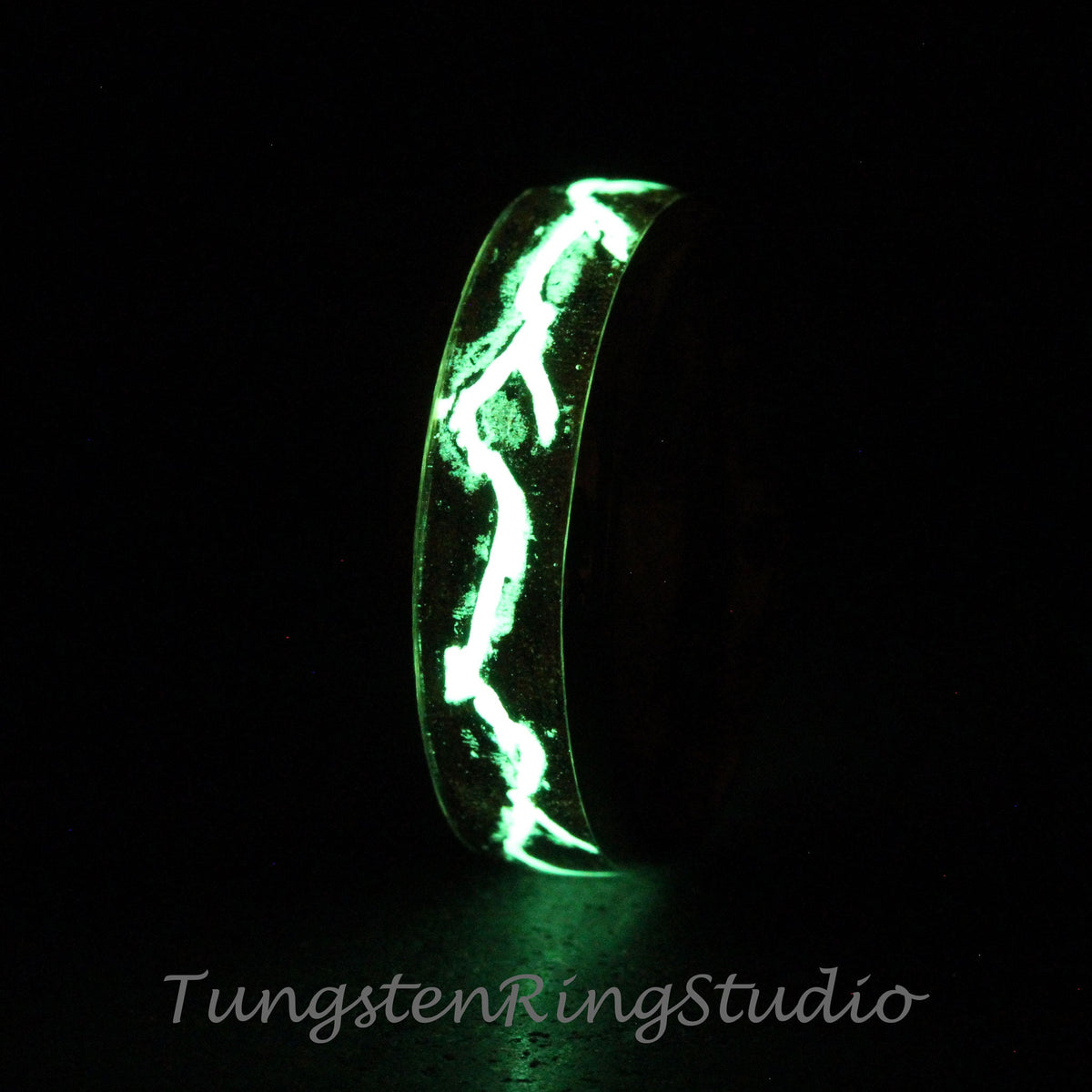 May include: A tungsten ring with a green glow-in-the-dark design. The design is a series of lines that resemble lightning bolts. The ring is shown in a dark setting.
