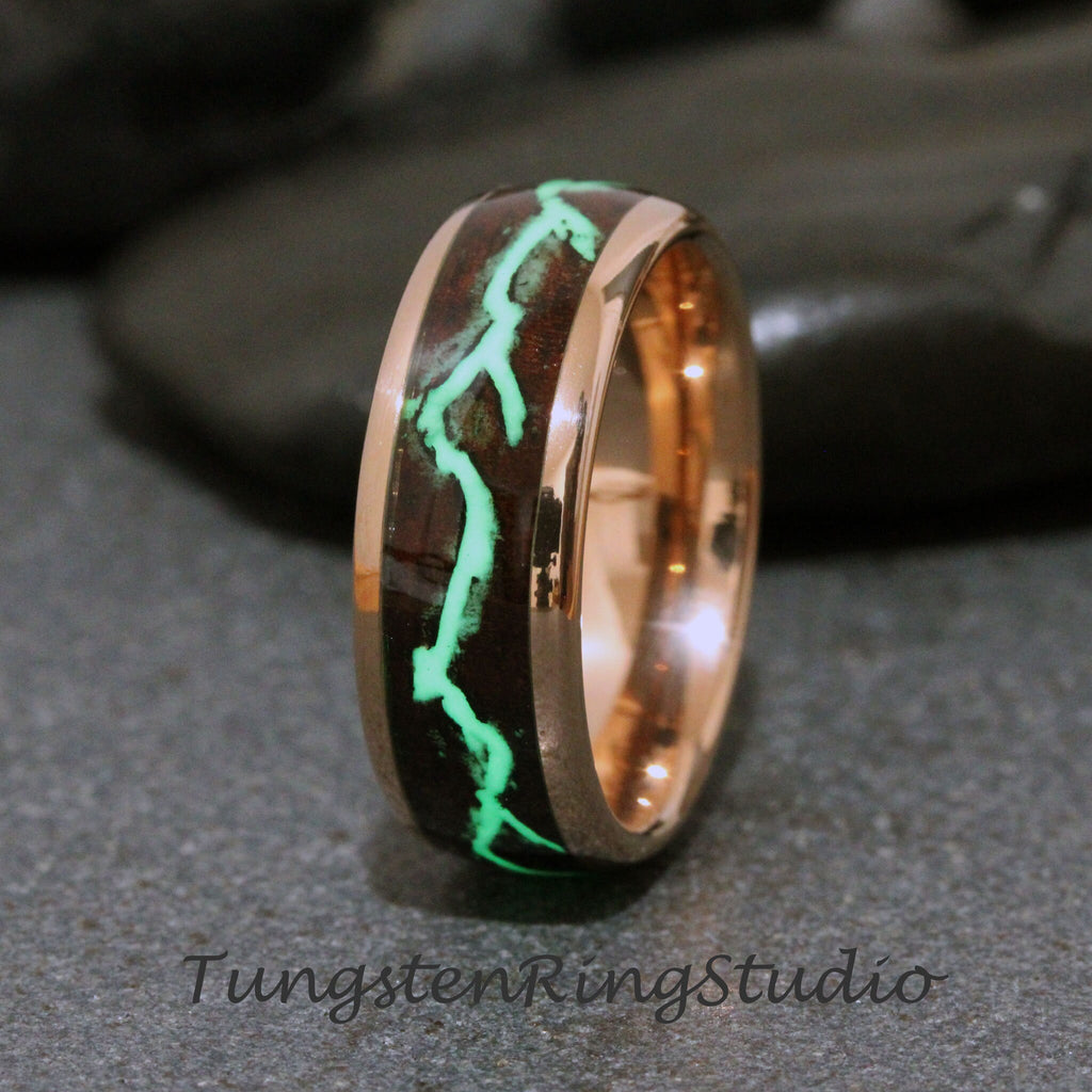 May include: A rose gold tungsten carbide ring with a dark brown wood inlay and a green glow-in-the-dark design. The ring is a classic style with a beveled edge.