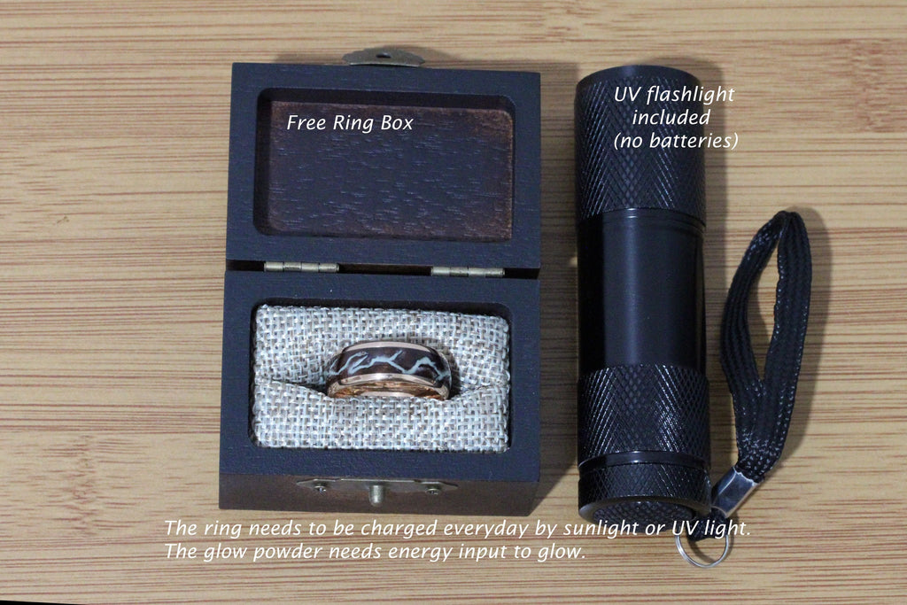 May include: A black wooden ring box with a brown velvet interior, containing a glow-in-the-dark ring with a black and white design. A black UV flashlight with a black strap is included. The text "Free Ring Box" and "UV flashlight included (no batteries)" is visible on the image. The text "The ring needs to be charged everyday by sunlight or UV light. The glow powder needs energy input to glow." is also visible.