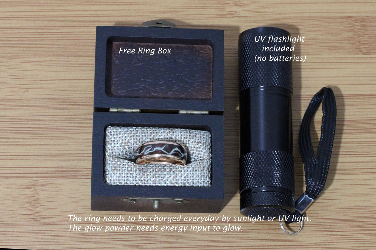 May include: A black wooden ring box with a brown velvet interior, containing a glow-in-the-dark ring with a black and white design. A black UV flashlight with a black strap is included. The text "Free Ring Box" and "UV flashlight included (no batteries)" is visible on the image. The text "The ring needs to be charged everyday by sunlight or UV light. The glow powder needs energy input to glow." is also visible.