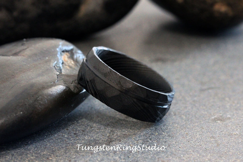 May include: A black tungsten carbide ring with a brushed finish and a single, thin, horizontal groove. The ring is on a gray surface.