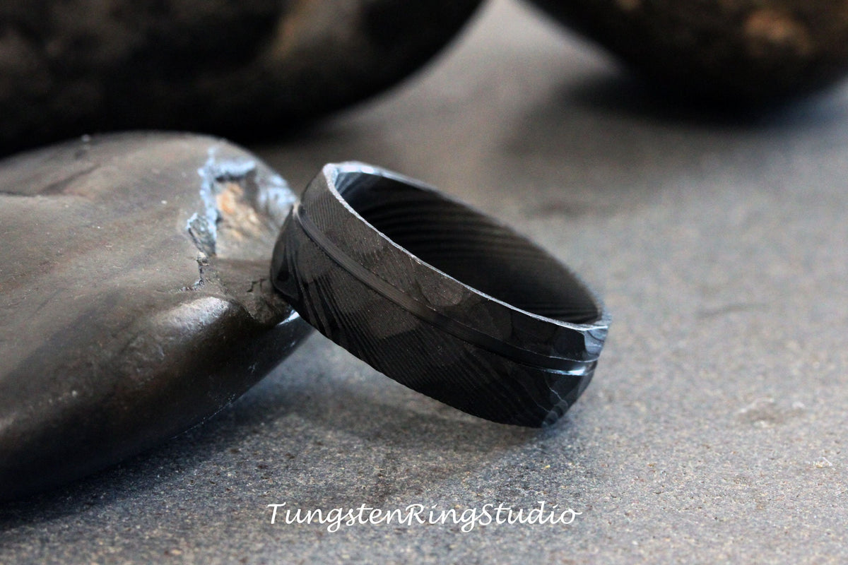 May include: A black tungsten carbide ring with a brushed finish and a single, thin, horizontal groove. The ring is on a gray surface.