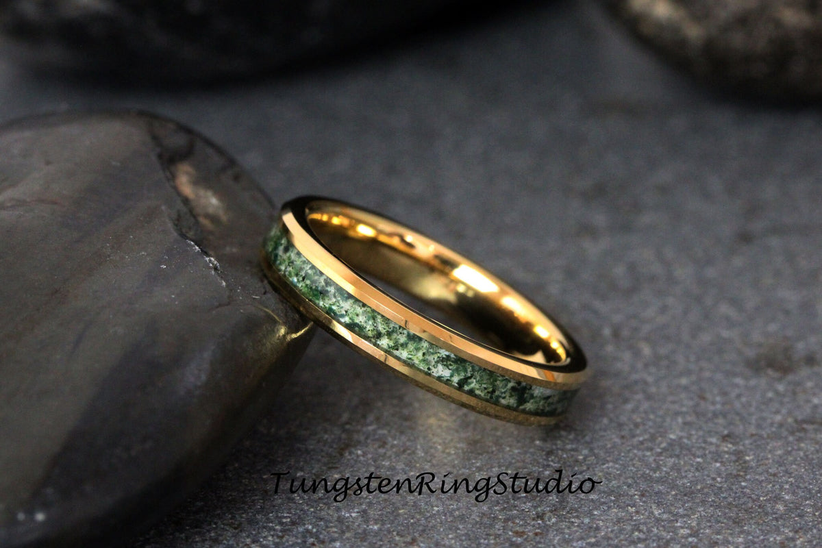 May include: A gold tungsten ring with a green and white inlay. The ring is on a dark gray rock.