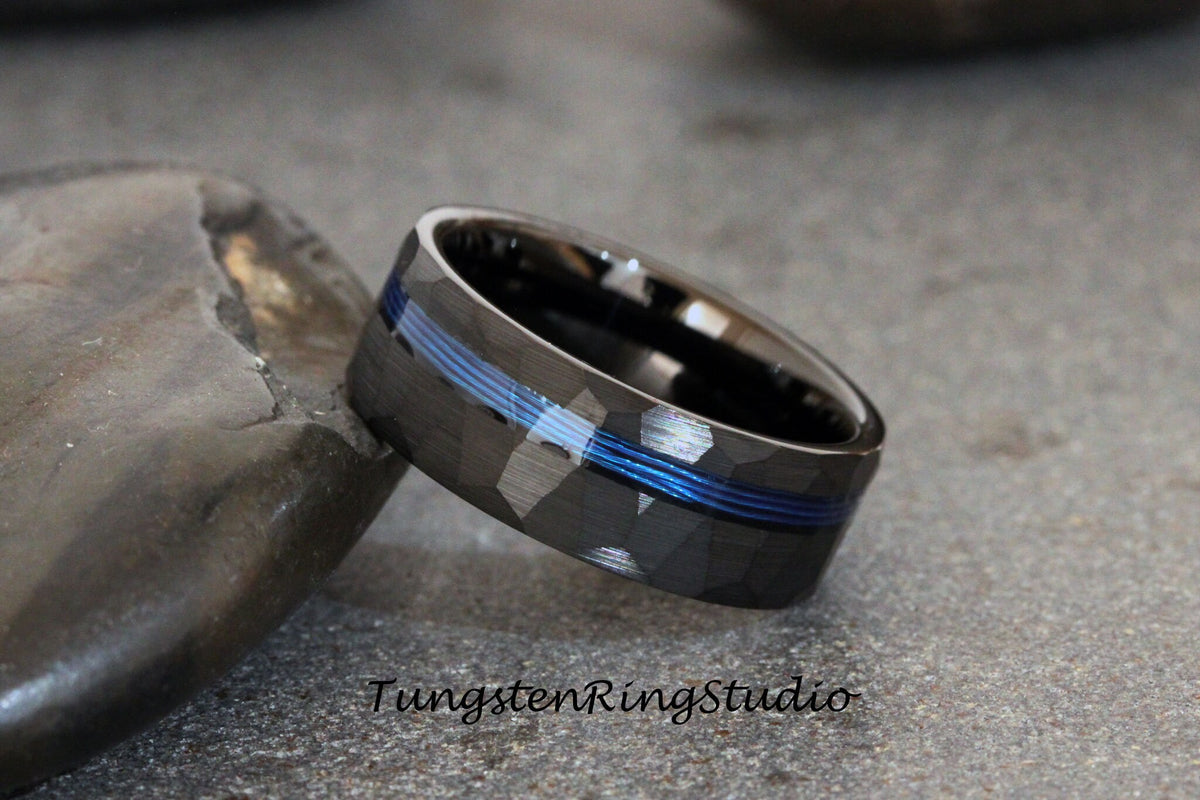 May include: A black tungsten carbide ring with a hammered finish and a blue line inlay. TungstenRingStudio is printed below the ring.