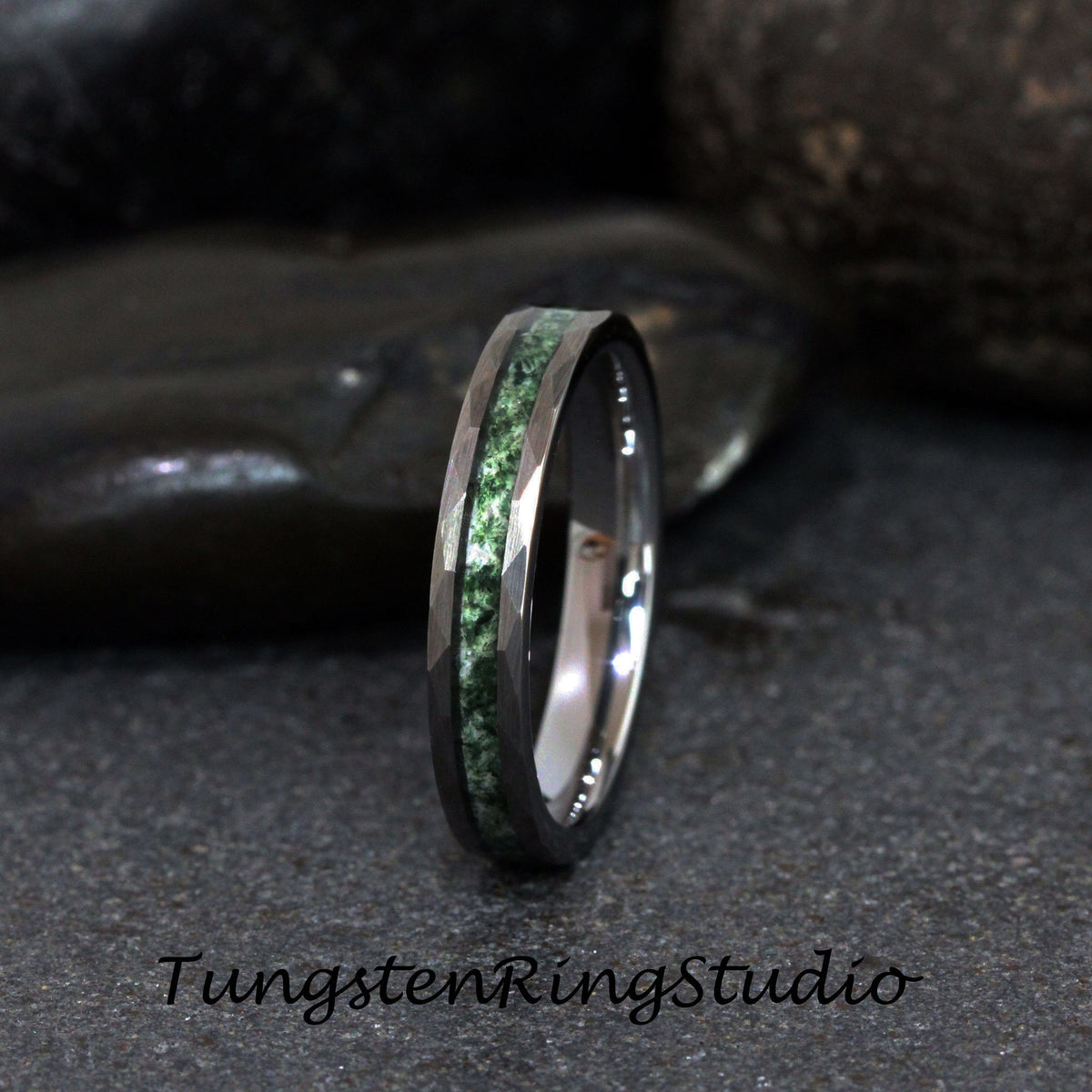 May include: A tungsten ring with a faceted, dark gray band and a central inlay of green stone. The ring is set against a dark gray background. The text "TungstenRingStudio" is at the bottom.
