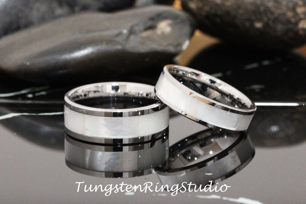 May include: Two silver tungsten carbide rings with a white mother of pearl inlay. The rings are on a black surface.
