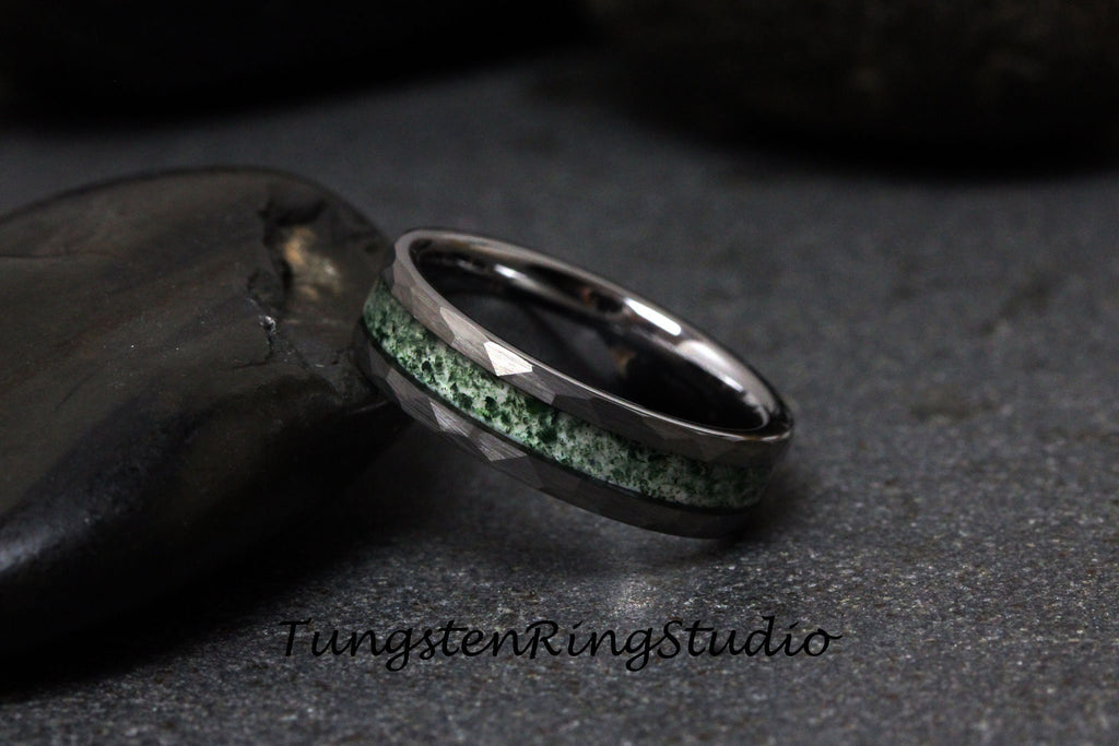 May include: A dark gray tungsten ring with a hammered finish and a green stone inlay. The ring is set against a dark background, highlighting its metallic sheen and textured design. The text "TungstenRingStudio" is visible.