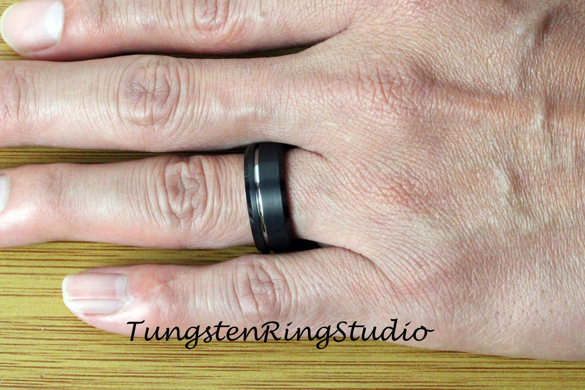 May include: A black tungsten ring with a silver inlay. The ring is on a person's finger.