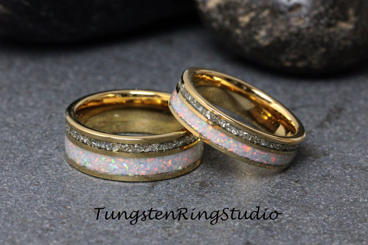May include: Two gold tungsten carbide rings with white opal and diamond inlay. The rings are on a gray surface.