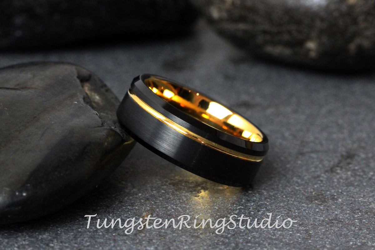 May include: A black tungsten carbide ring with a gold inlay. The ring has a brushed finish and a beveled edge.