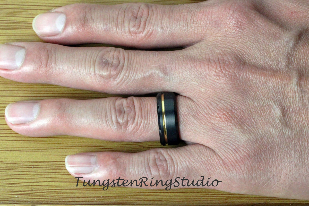 May include: A black tungsten carbide ring with a gold inlay. The ring is worn on a person's finger.
